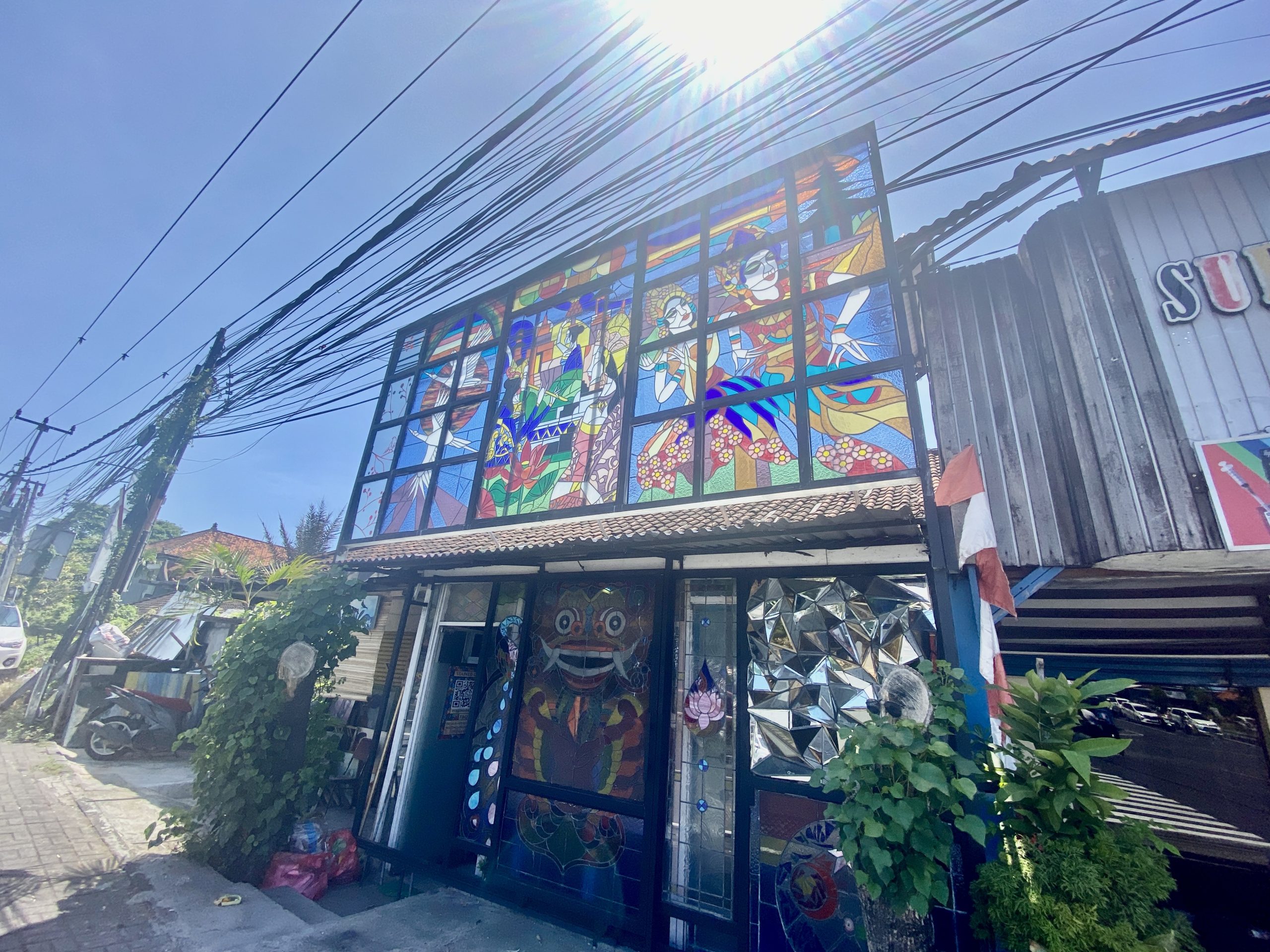 Front of a stained glass workshop in Sanur, Bali, with a large hand-made leaded glass window of Balinese legong dancers above a doorway decorated with a Barong mosaic