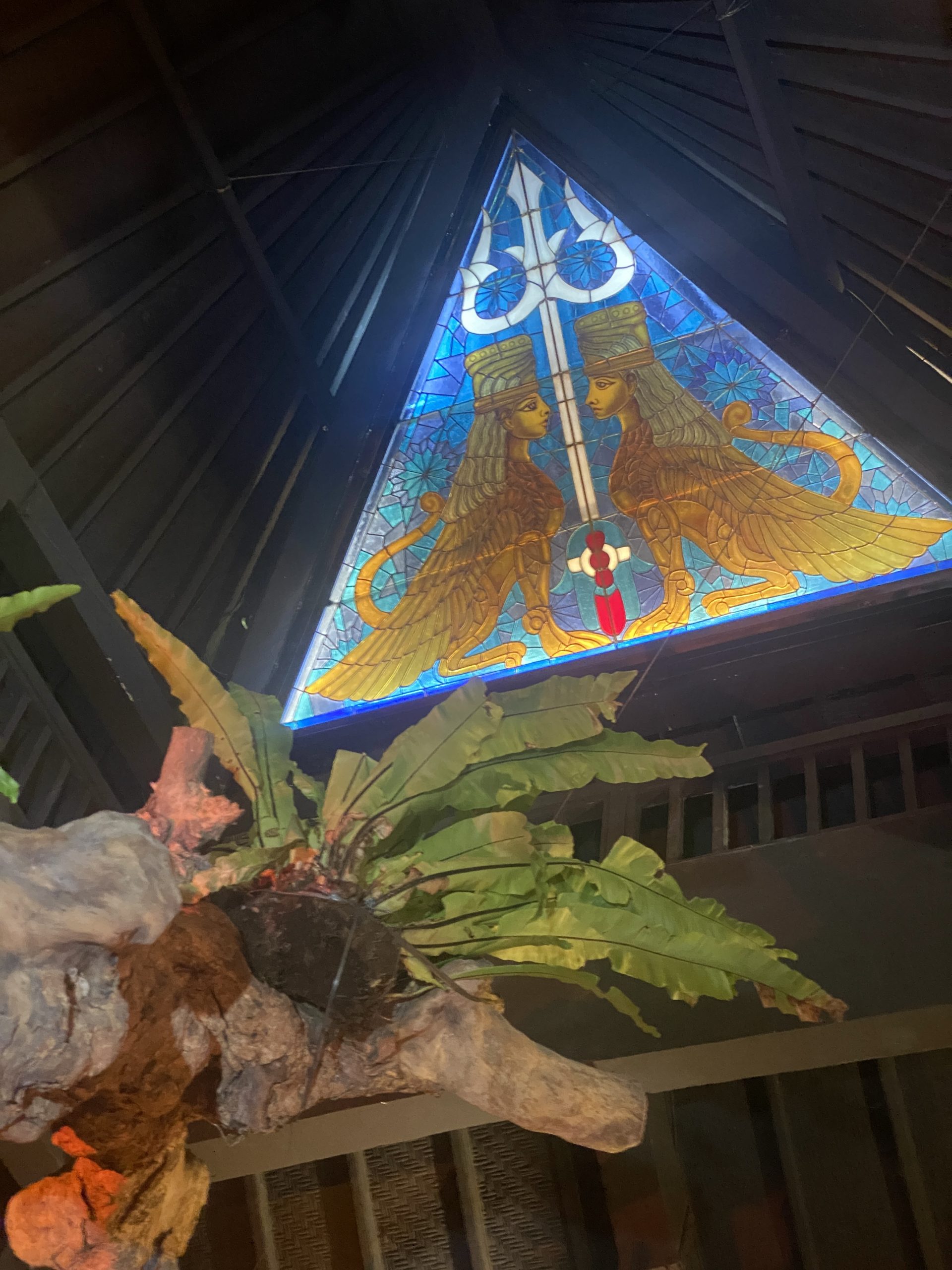 Backlit stained glass ceiling panel at Zest Ubud showing two mirrored winged guardian figures in amber and gold against a fractured turquoise ground.
